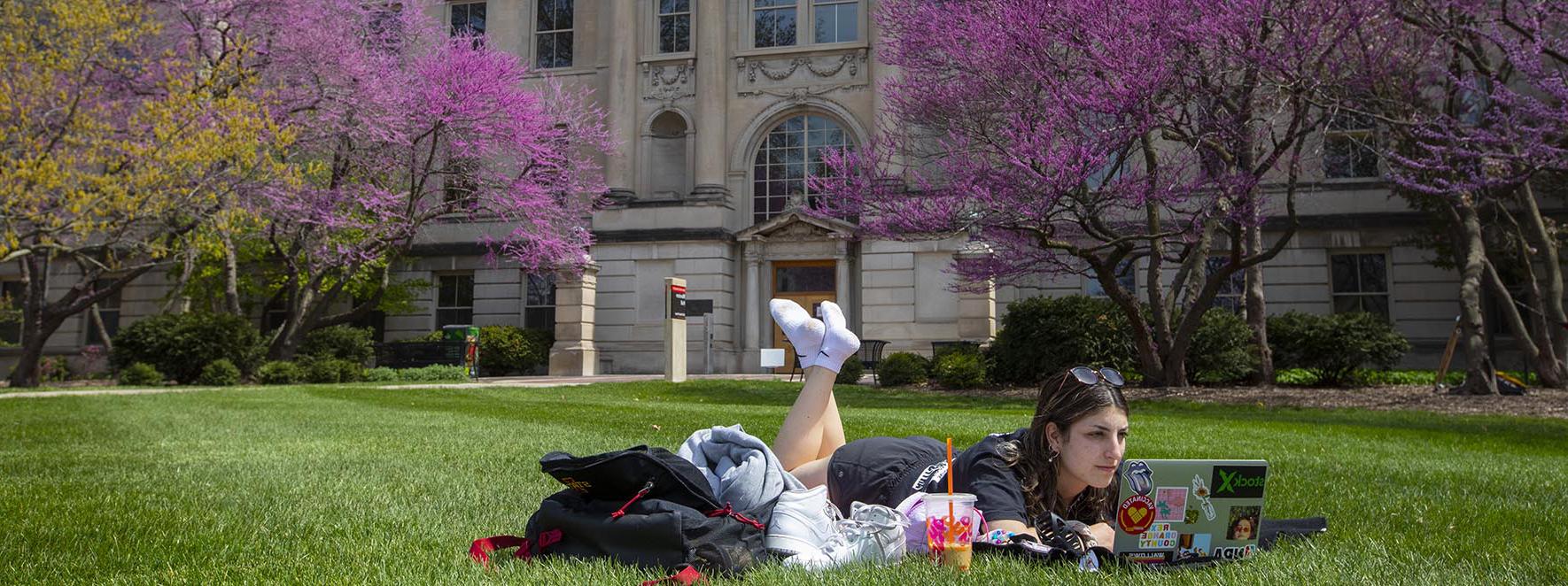 一名学生躺在马斯顿大厅外的草地上摆弄电脑